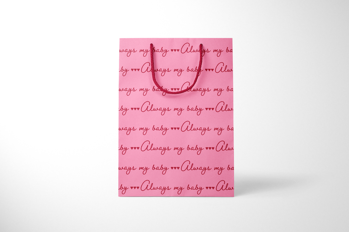 Front view of bright pink gift bag with repeating red script text that reads ‘Always my baby’ with small heart accents, finished with matching red rope handles.