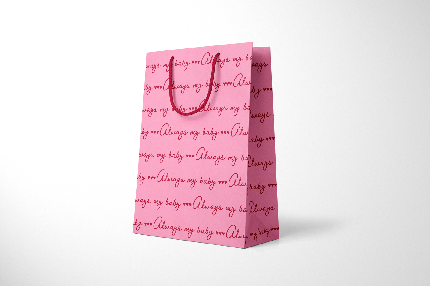 Side view of bright pink gift bag with repeating red script text that reads ‘Always my baby’ with small heart accents, finished with matching red rope handles.