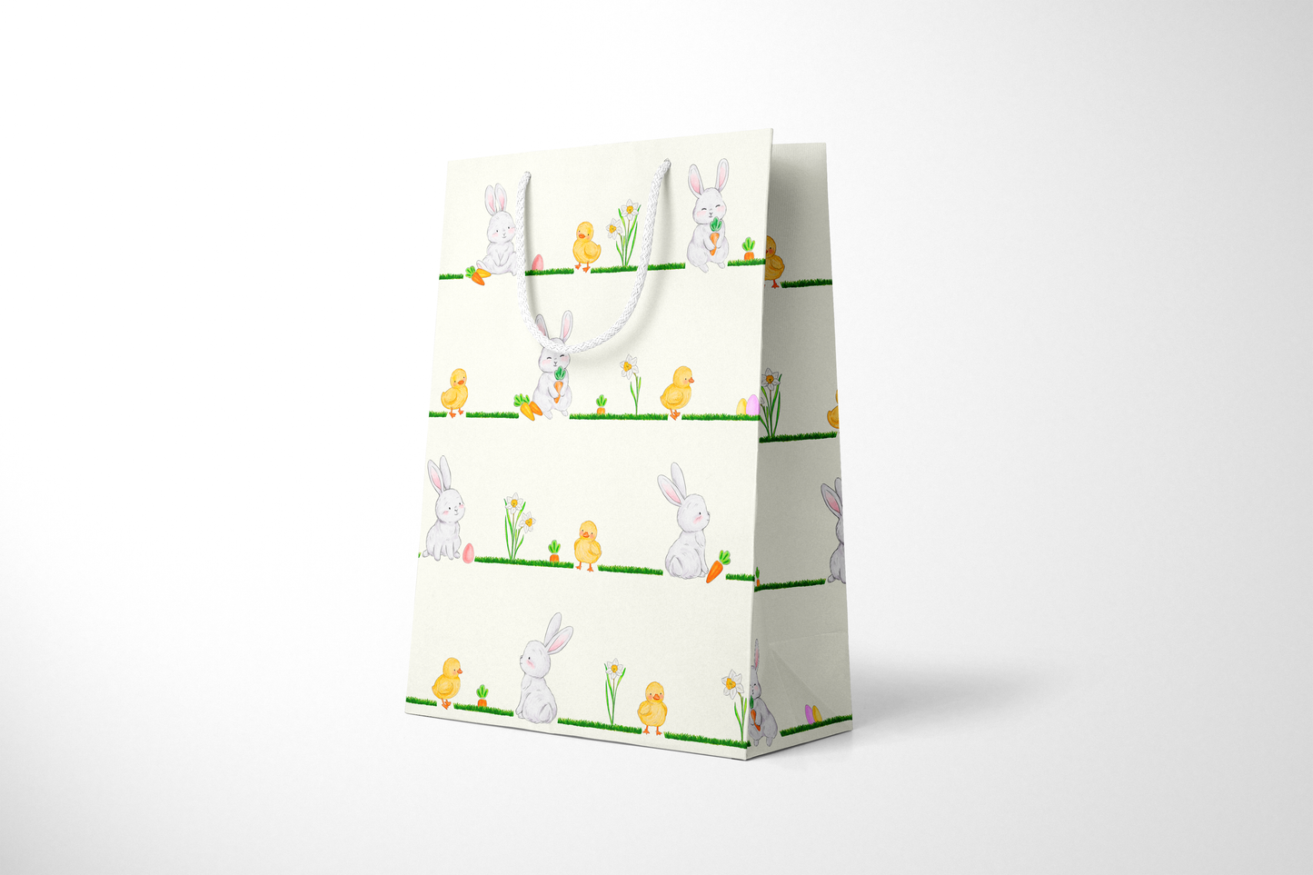 Side view of cream Easter gift bag featuring a repeating horizontal pattern of watercolor-style bunnies, ducklings, daffodils, carrots, and Easter eggs on grassy lines, with a white rope handle.