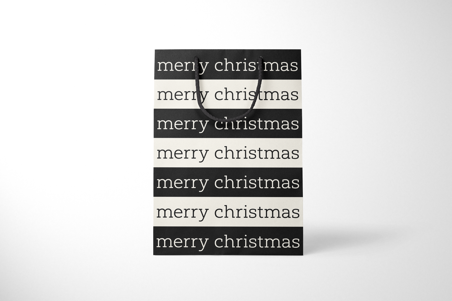 Front view of modern minimalist gift bag featuring alternating black and cream stripes with repeating ‘merry christmas’ text in bold typography, finished with sturdy black rope handles.