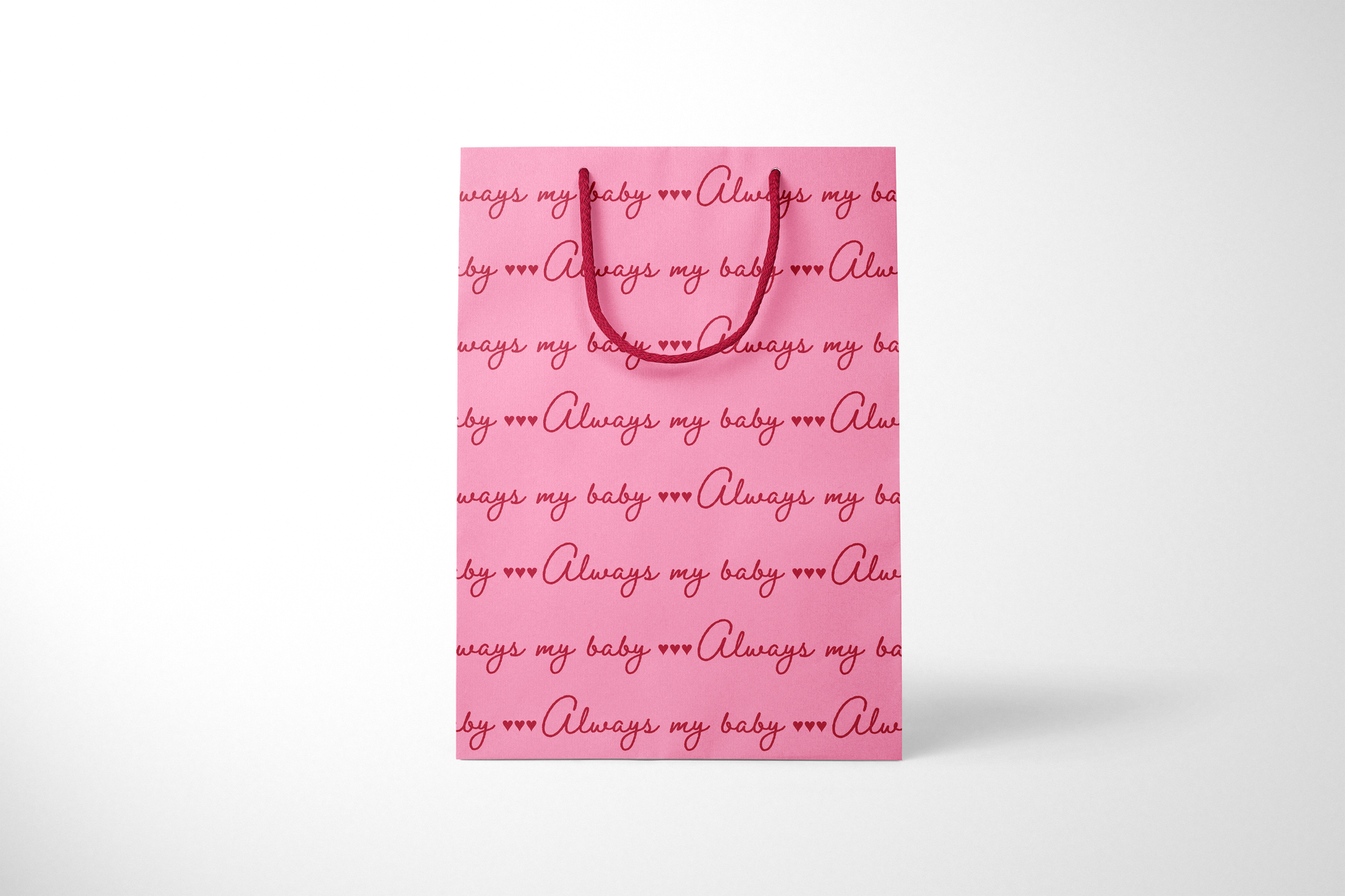 Front view of bright pink gift bag with repeating red script text that reads ‘Always my baby’ with small heart accents, finished with matching red rope handles.
