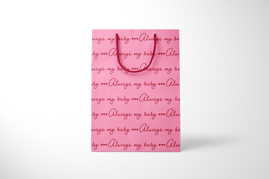 Front view of bright pink gift bag with repeating red script text that reads ‘Always my baby’ with small heart accents, finished with matching red rope handles.
