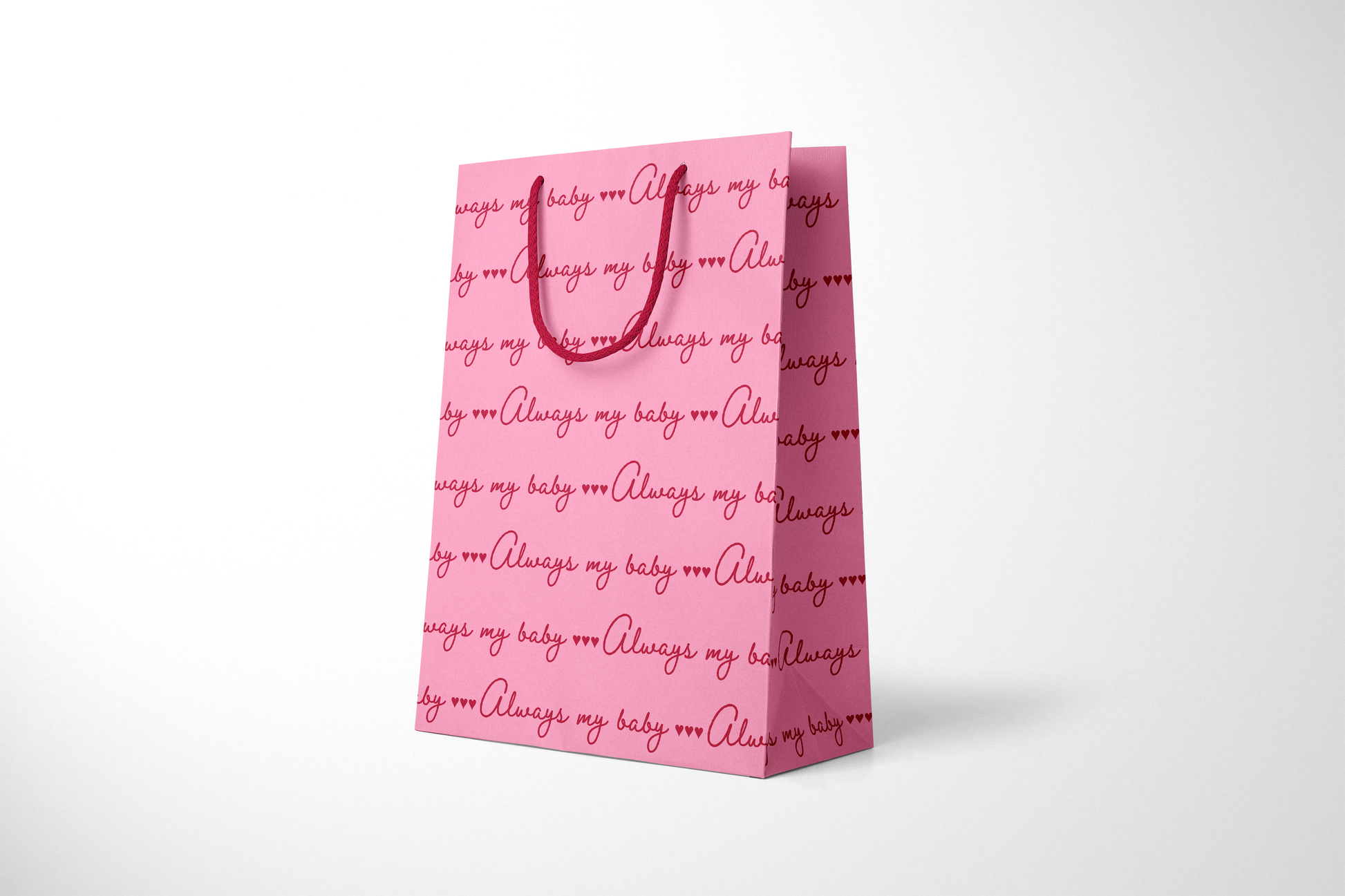 Side view of bright pink gift bag with repeating red script text that reads ‘Always my baby’ with small heart accents, finished with matching red rope handles.