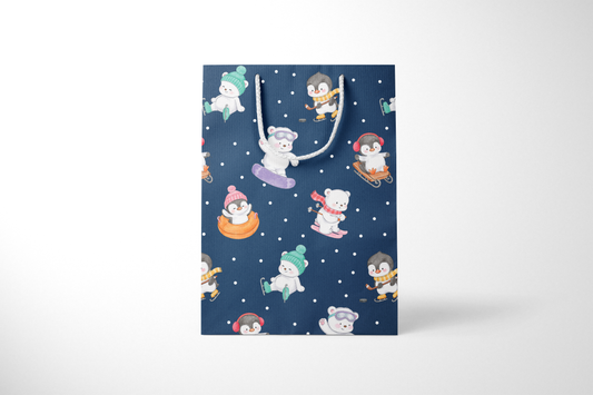 Front view of a navy blue gift bag with playful illustrations of polar bears and penguins enjoying winter activities like snowboarding, sledding, tubing, skating, and hockey, accented with white snow dots and white rope handles.