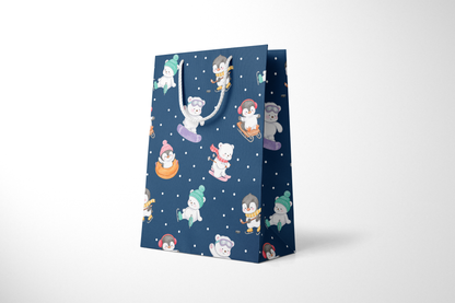 Side view of a navy blue gift bag with playful illustrations of polar bears and penguins enjoying winter activities like snowboarding, sledding, tubing, skating, and hockey, accented with white snow dots and white rope handles.