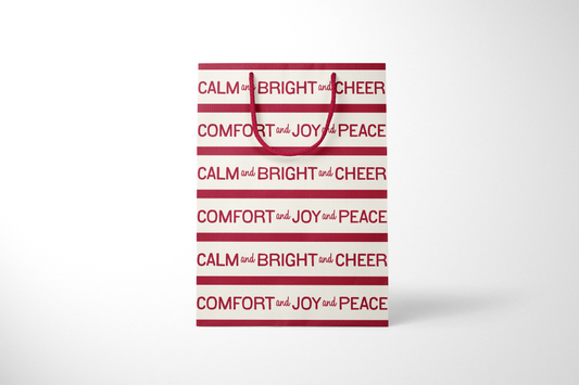 Front view of modern holiday gift bag with alternating red and cream stripes featuring repeating typographic phrases ‘CALM and BRIGHT and CHEER’ and ‘COMFORT and JOY and PEACE,’ finished with matching red rope handles.