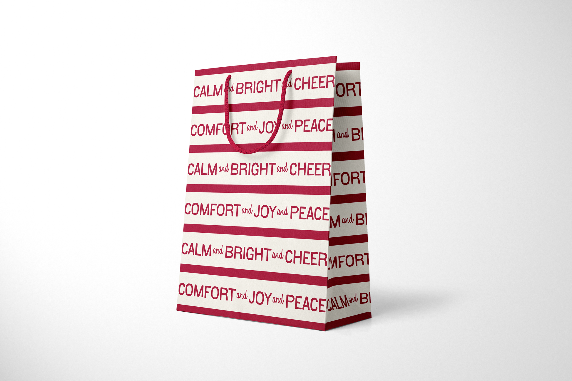 Side view of modern holiday gift bag with alternating red and cream stripes featuring repeating typographic phrases ‘CALM and BRIGHT and CHEER’ and ‘COMFORT and JOY and PEACE,’ finished with matching red rope handles.