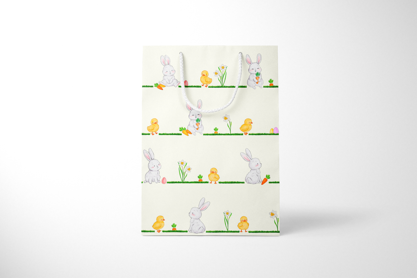 Front view of cream Easter gift bag featuring a repeating horizontal pattern of watercolor-style bunnies, ducklings, daffodils, carrots, and Easter eggs on grassy lines, with a white rope handle.