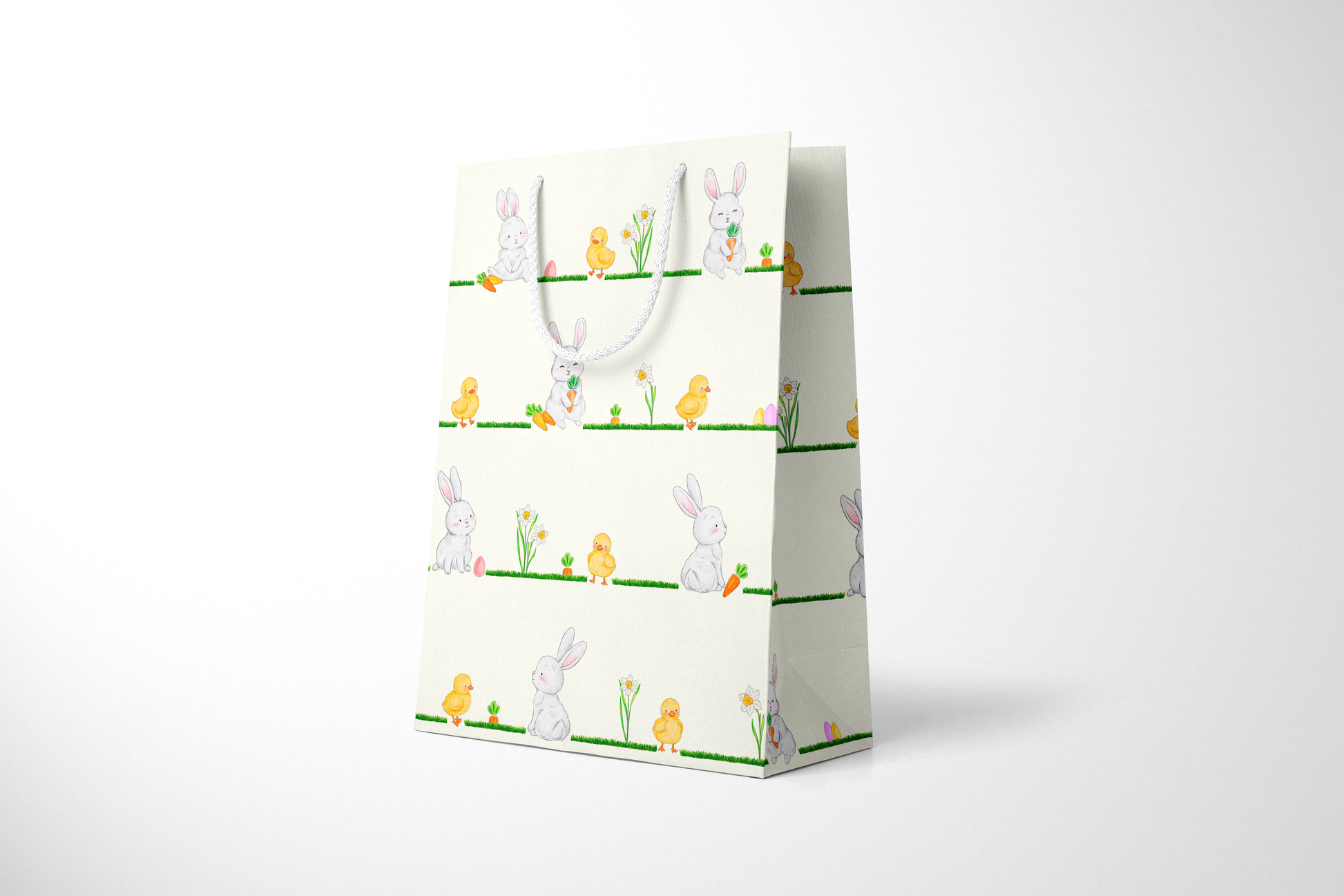 Side view of cream Easter gift bag featuring a repeating horizontal pattern of watercolor-style bunnies, ducklings, daffodils, carrots, and Easter eggs on grassy lines, with a white rope handle.