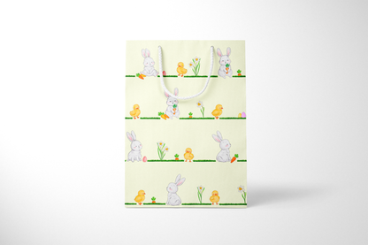Front view of light yellow Easter gift bag featuring a repeating horizontal pattern of watercolor-style bunnies, ducklings, daffodils, carrots, and Easter eggs on grassy lines, with a white rope handle.