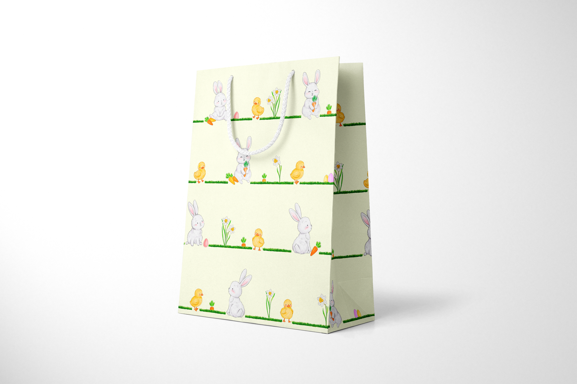 Side view of light yellow Easter gift bag featuring a repeating horizontal pattern of watercolor-style bunnies, ducklings, daffodils, carrots, and Easter eggs on grassy lines, with a white rope handle.