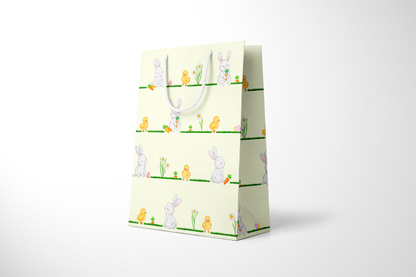 Side view of light yellow Easter gift bag featuring a repeating horizontal pattern of watercolor-style bunnies, ducklings, daffodils, carrots, and Easter eggs on grassy lines, with a white rope handle.