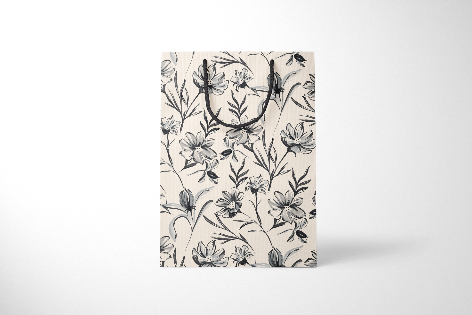 Front view of cream gift bag with hand-drawn black and gray floral illustrations, finished with black rope handles.
