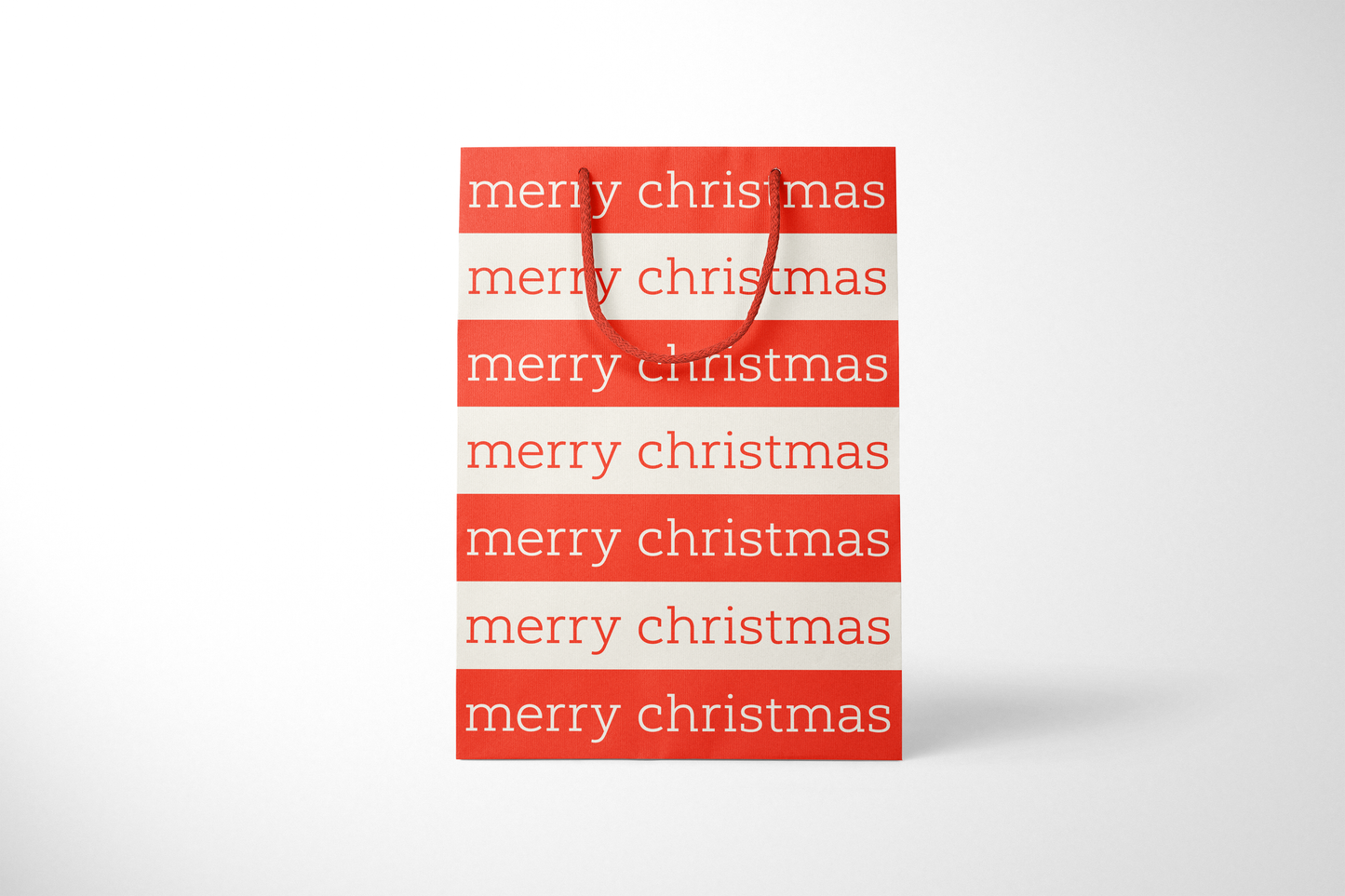 Front view of modern minimalist gift bag featuring alternating red and cream stripes with repeating ‘merry christmas’ text in bold typography, finished with sturdy red rope handles.