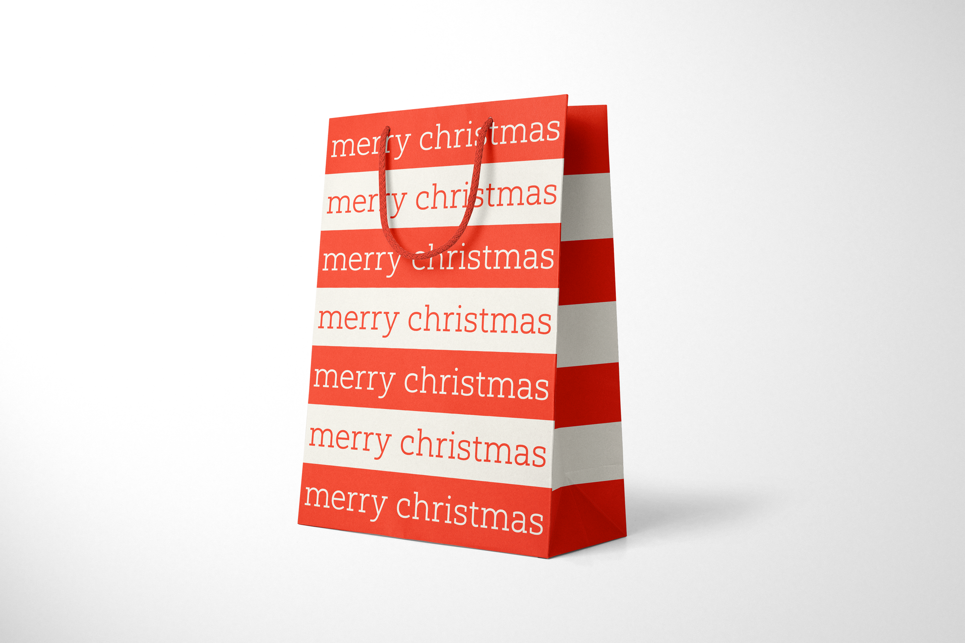 Side view of modern minimalist gift bag featuring alternating red and cream stripes with repeating ‘merry christmas’ text in bold typography, finished with sturdy red rope handles.