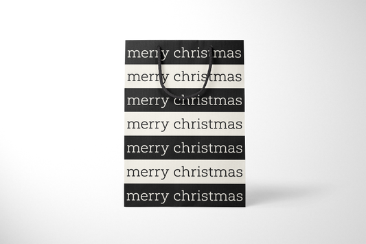 Front view of modern minimalist gift bag featuring alternating black and cream stripes with repeating ‘merry christmas’ text in bold typography, finished with sturdy black rope handles.