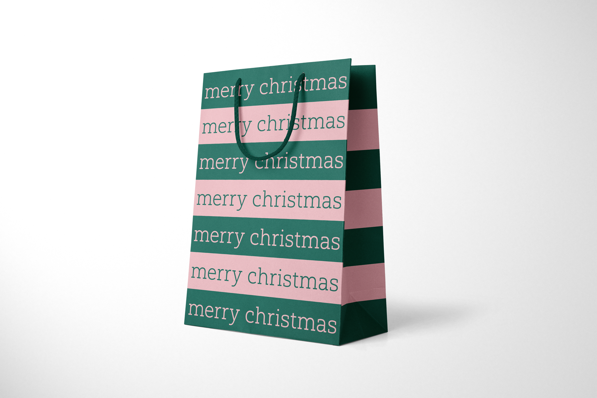 Side view of modern minimalist gift bag featuring alternating pink and green stripes with repeating ‘merry christmas’ text in bold typography, finished with sturdy dark green rope handles.