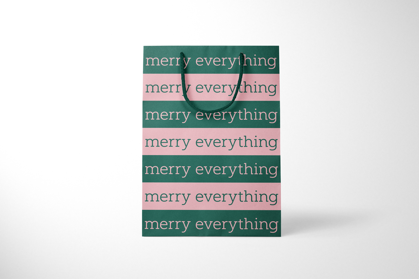 Front view of modern minimalist gift bag featuring alternating pink and green stripes with repeating ‘merry everything' text in bold typography, finished with sturdy dark green rope handles.