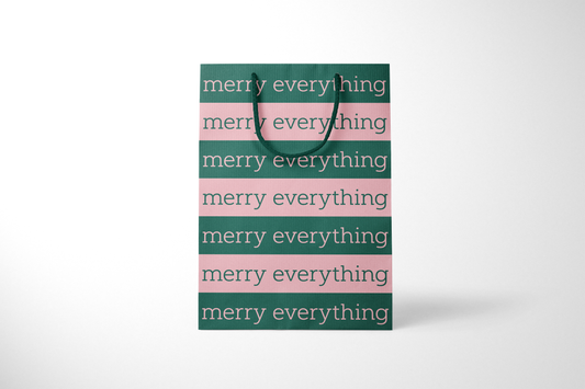 Front view of modern minimalist gift bag featuring alternating pink and green stripes with repeating ‘merry everything' text in bold typography, finished with sturdy dark green rope handles.