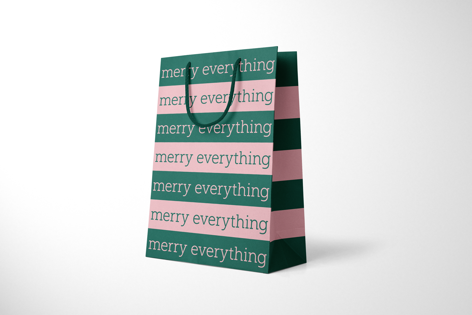 Side view of modern minimalist gift bag featuring alternating pink and green stripes with repeating ‘merry everything' text in bold typography, finished with sturdy dark green rope handles.