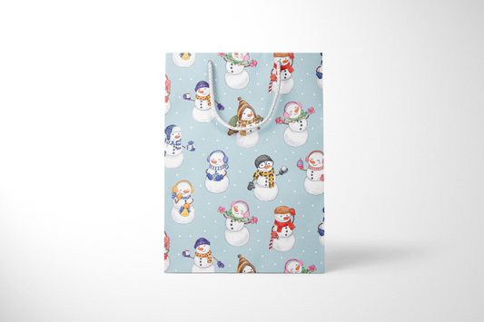 Front view of light blue gift bag with a whimsical snowman pattern, featuring snowmen in colorful scarves, hats, and earmuffs, holding holiday items like gifts, candy canes, and hot cocoa, with white rope handles and scattered snow dots and a white rope handles.