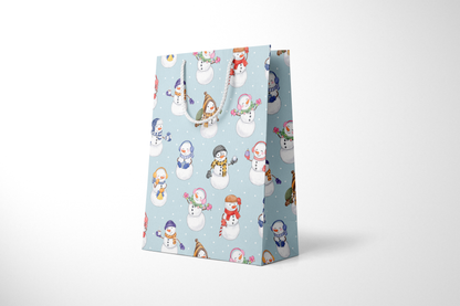 Side view of light blue gift bag with a whimsical snowman pattern, featuring snowmen in colorful scarves, hats, and earmuffs, holding holiday items like gifts, candy canes, and hot cocoa, with white rope handles and scattered snow dots and a white rope handles.