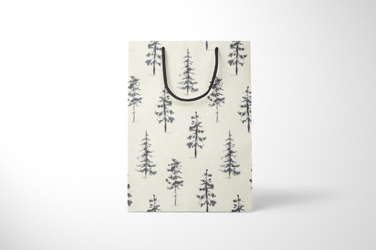 Front view of cream-colored holiday gift bag with hand-painted black and gray evergreen trees scattered across the surface, accented with falling snow dots, and finished with black rope handles.